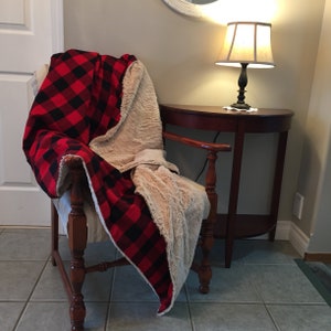 May include: A red and black buffalo plaid blanket draped over a wooden chair. The blanket has a soft, cream-colored sherpa lining. The chair is positioned next to a small, dark wooden side table with a lamp.
