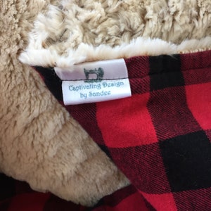 May include: A close-up of a blanket with a red and black plaid pattern. The blanket has a soft, beige faux fur trim. A label reads "Captivating Design by Sandee" with a sewing machine illustration.