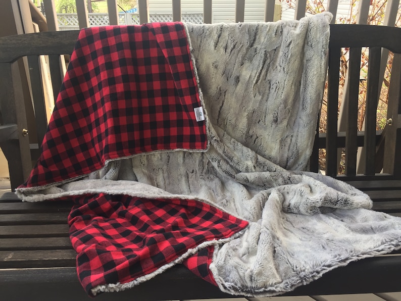 May include: A red and black plaid blanket with a soft, gray faux fur side. The blanket is folded and draped over a wooden bench.
