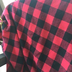 May include: Red and black buffalo plaid fabric. The fabric is folded over a dark surface, showing the pattern of large, square checks. The material appears to be soft and possibly flannel.