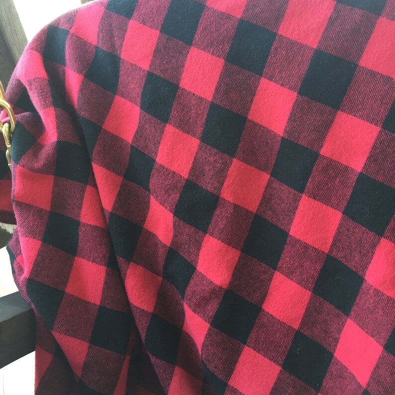 May include: Red and black buffalo plaid fabric. The fabric is folded over a dark surface, showing the pattern of large, square checks. The material appears to be soft and possibly flannel.