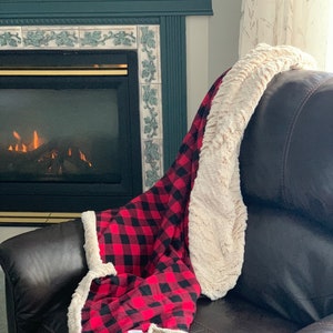 May include: A red and black buffalo plaid blanket with a cream-colored sherpa lining is draped over a brown leather armchair. The blanket is folded, showcasing the plaid pattern and the soft texture of the sherpa.