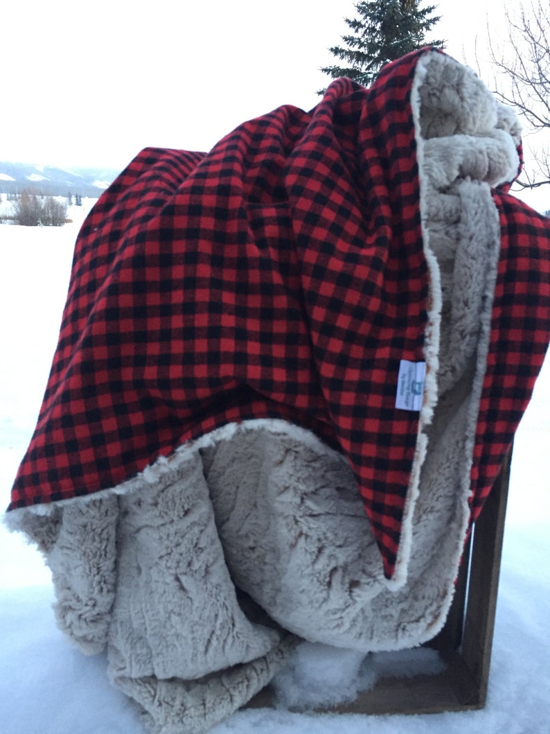 May include: A red and black buffalo plaid blanket with a soft, gray faux fur lining. The blanket is folded over a wooden crate in a snowy outdoor setting. The blanket is a cozy home accessory.