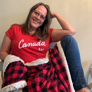 May include: A woman wearing a red t-shirt with the text "Canada GAP" in white lettering, sitting on a white bench with a red and black plaid blanket.