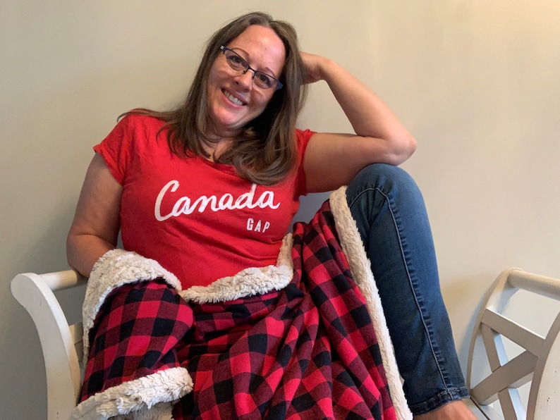 May include: A woman wearing a red t-shirt with the text "Canada GAP" in white lettering, sitting on a white bench with a red and black plaid blanket.