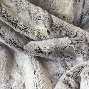 May include: Close-up of a soft, plush faux fur fabric in a gray and white color scheme. The fabric has a long pile and a subtle striped pattern.