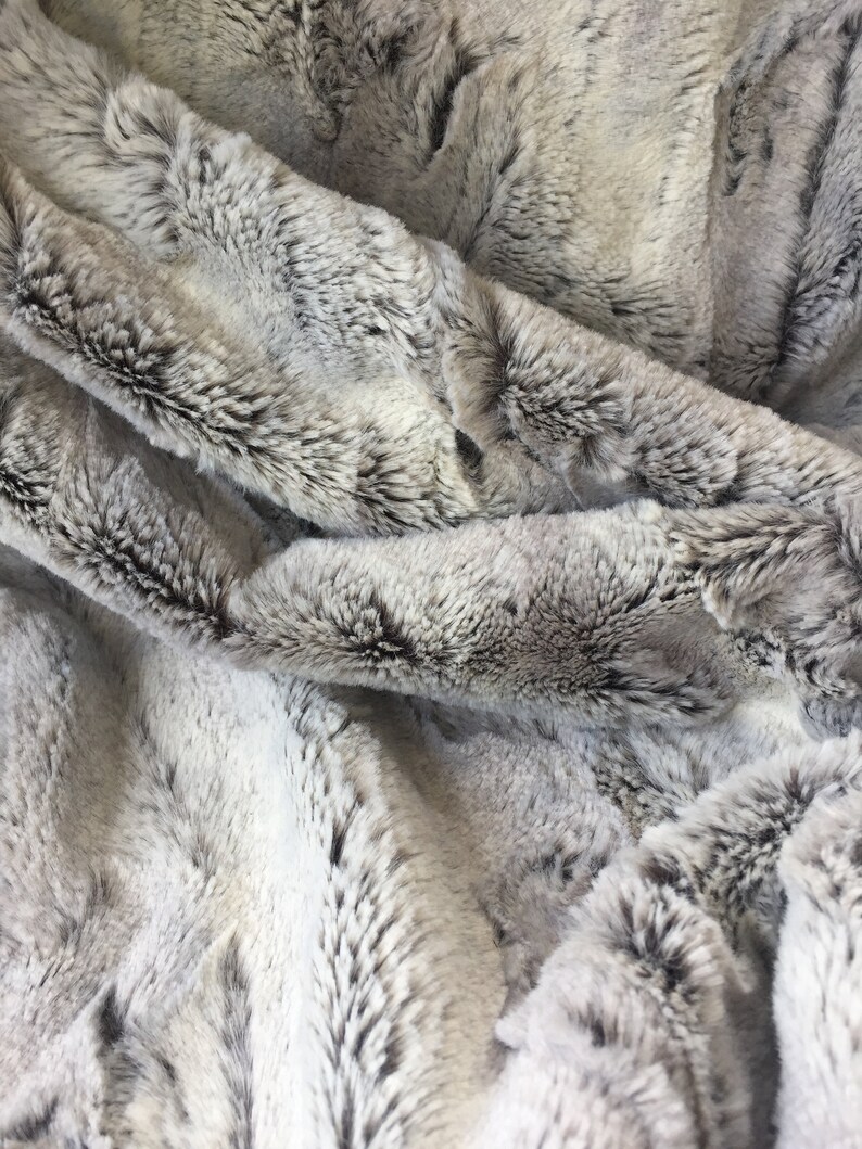 May include: Close-up of a soft, plush faux fur fabric in a gray and white color scheme. The fabric has a long pile and a subtle striped pattern.