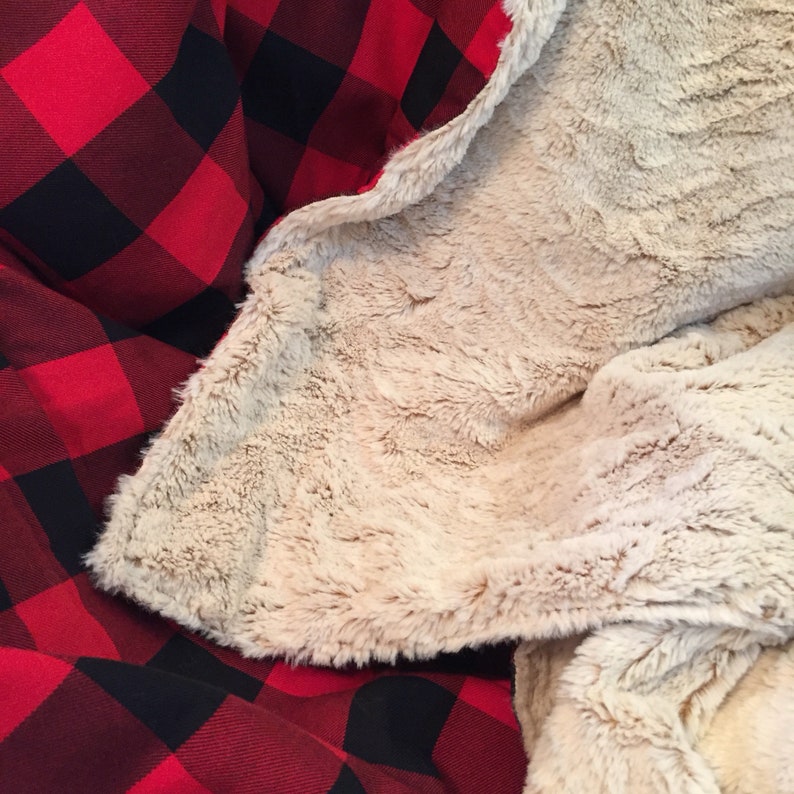 May include: A cozy blanket with a red and black buffalo plaid pattern on one side and a soft, tan faux fur on the other. The blanket is folded, showcasing the contrasting textures and colors.