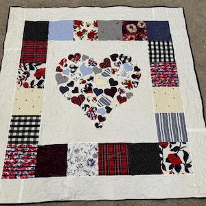 May include: A handmade quilt featuring a heart design at the centre. The heart is made from various patterned fabrics, including stripes, florals, and plaids. The quilt's border has a patchwork design with similar patterns. The background is white.