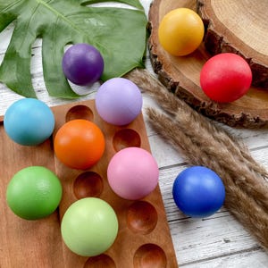 May include: A set of ten wooden balls in various colours, including red, orange, yellow, green, blue, purple, and pink. The balls are arranged in a wooden tray with individual compartments.