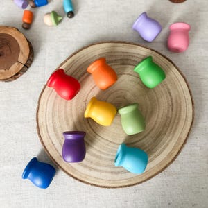 May include: A selection of brightly coloured wooden toy cups in red, orange, yellow, green, blue, purple, and pink, displayed on a wooden slice. Other wooden toys, including acorn shapes, are scattered around. These toys are designed for children.