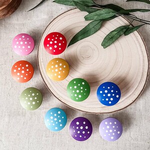 May include: Nine colorful wooden knobs with white polka dots. The knobs are arranged in a circle on a wooden surface. The knobs are in the colors of the rainbow, starting with red and ending with purple.