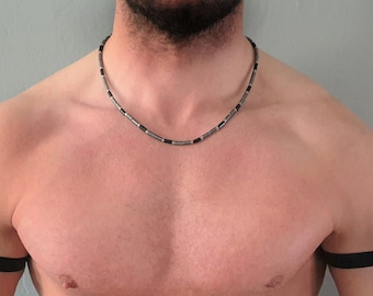 May include: A necklace featuring alternating cylindrical silver-toned metal beads and small black beads, paired with two plain black elastic armbands worn around the upper arms. The necklace rests at the base of the neck, while the armbands are positioned symmetrically on the biceps.