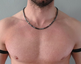 May include: A necklace composed of small, dark, square-shaped metallic beads worn around the neck, paired with two plain black elastic bands positioned around each upper arm. The necklace features a uniform, repeating pattern of dark, reflective beads.