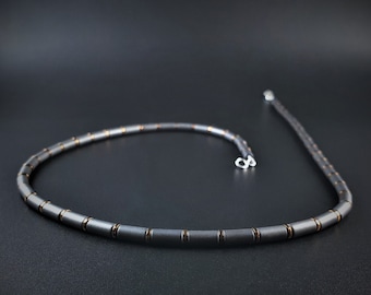 May include: A necklace composed of alternating dark gray cylindrical beads and thin copper-colored spacers. The strand features a silver-toned metal clasp at the end, creating a continuous circular arrangement of uniform, matte-finished components.