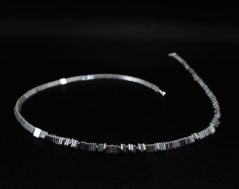 May include: A necklace composed of numerous small, dark gray, rectangular metallic beads arranged in a continuous strand. The design features evenly spaced, smooth, square silver-toned metal accents interspersed among the smaller dark beads.