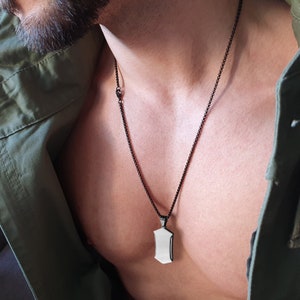 Könnte beinhalten: Eine schwarze Anhängerhalskette mit einem rechteckigen, schildförmigen Anhänger. Der Anhänger hat eine silberfarbene, reflektierende Oberfläche. Die Halskette hat eine dünne, schwarze Kugelkette. Im Hintergrund eine grüne Jacke.