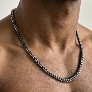 May include: A silver-toned metal chain necklace featuring a series of interlocking, textured links. The chain rests around the neck, displaying a consistent, repeating pattern of small, angled segments that create a subtle, darkened finish.