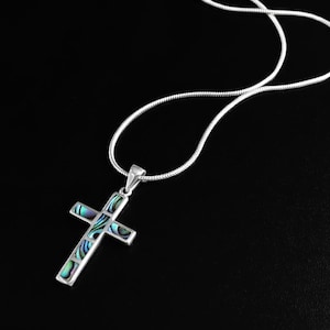 Può includere: Collana con pendente a croce in argento con intarsio in abalone. La croce è sospesa a una delicata catena d'argento. L'intarsio in abalone presenta iridescenze blu, verdi e viola. La collana è esposta su sfondo nero.