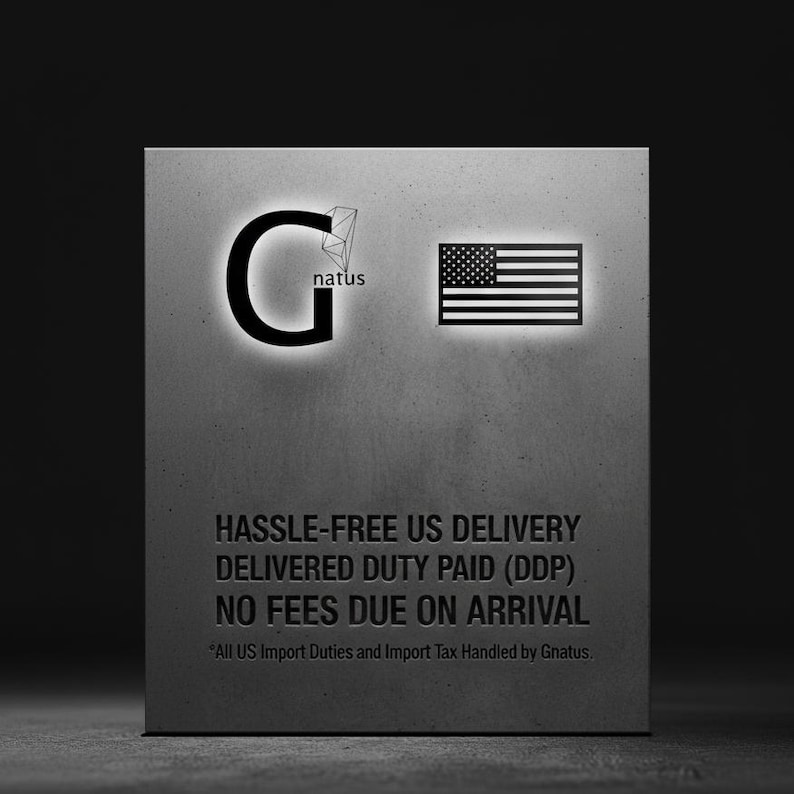 May include: A rectangular gray sign featuring the black letter G with a geometric diamond icon and the text natus, alongside a black and white American flag. Below, the text reads HASSLE-FREE US DELIVERY DELIVERED DUTY PAID (DDP) NO FEES DUE ON ARRIVAL.