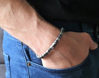 May include: A bracelet made of black and silver square beads. The bracelet is worn on a person's wrist.