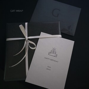 May include: A black square gift box wrapped with a thin white ribbon, accompanied by a light-colored card featuring a line-art illustration of a gift box with a heart, the text HAPPY BIRTHDAY, and the name Steve, plus a dark envelope with a G logo.