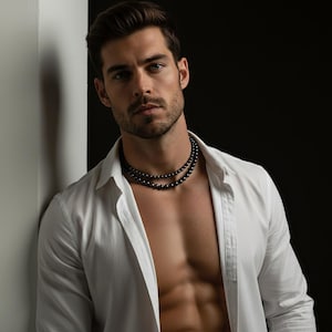 Puede incluir: Un collar de cuentas negras de dos hilos. El collar se lleva sobre una camisa blanca abotonada. El fondo es blanco y negro. El collar es una joya.