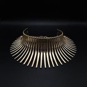 Könnte beinhalten: Eine goldfarbene Statement-Halskette mit Kragendesign. Die Halskette hat zahlreiche vertikale Stäbe, die sich vom Kragen ausfächern und einen auffälligen Look erzeugen. Der Schmuck ist vor einem schwarzen Hintergrund platziert.