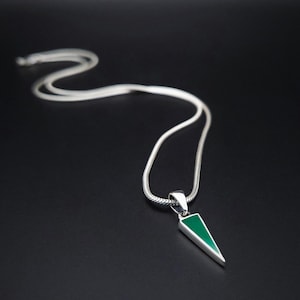 Puede incluir: Un collar de cadena de serpiente plateada con un colgante triangular que presenta una incrustación verde lisa y vibrante. El colgante está sujeto a la cadena mediante una reasa de plata pulida, creando una pieza de joyería elegante y definida.