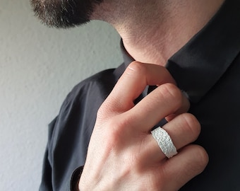 Könnte beinhalten: Ein silberner Ring mit einer strukturierten Oberfläche. Der Ring wird am Ringfinger einer Hand getragen.