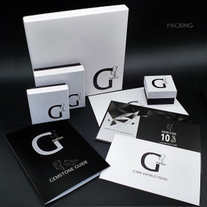 May include: A white box with the word "natus" and a stylized "G" logo. The box is surrounded by other smaller boxes and brochures. The brochures include information about gemstones, care instructions, and a certificate of authenticity. The text "PACKING" is visible in the upper right corner of the image.