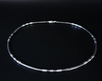 May include: A circular necklace composed of small, metallic, dark gray cylindrical beads arranged in a continuous strand. The necklace features a silver-toned metal lobster claw clasp at the back, connecting the two ends of the beaded chain securely.
