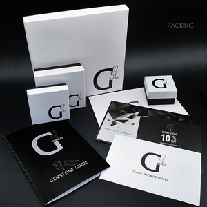 May include: A collection of white and black branded packaging boxes and printed cards featuring the G natus logo. The set includes a large square box, two smaller boxes, a Gemstone Guide, a Care Instructions card, and a Certificate of Authenticity.