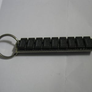 May include: A silver key chain with a black circuit board attached to it. The circuit board has a row of 16 black chips.