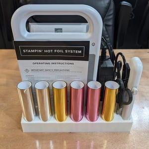 May include: A white Stampin' Hot Foil System machine with an operating instructions manual, a black power adapter, and six metallic foil rolls in silver, gold, rose gold, and pink colors, all organized in a white plastic base with a white tool.