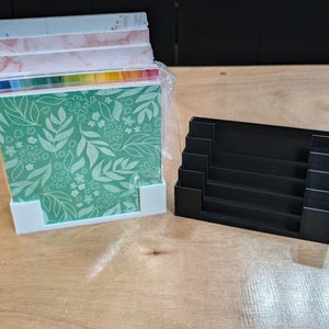 May include: A white plastic storage organizer with a stack of patterned paper inside. The organizer has a black plastic base with five slots for storing paper.