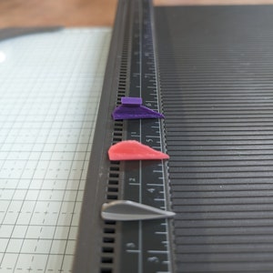 May include: Three small, 3D printed objects on a black ruler. The objects are a purple, a red, and a silver color. The ruler has markings in inches.