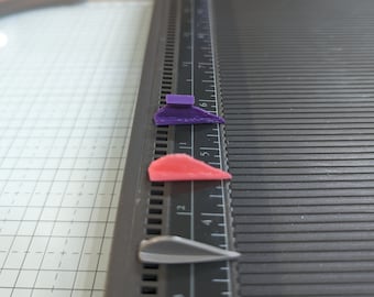 May include: Three small, 3D printed objects on a black ruler. The objects are purple, red, and silver. The ruler has markings in inches.