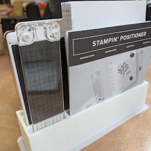 May include: A white plastic stamp positioner with a clear acrylic plate featuring a grid pattern. The set includes instructions and a clear acrylic guide. The text "STAMPIN' POSITIONER" is visible on the instructions.