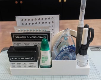 Pode incluir: Um organizador branco com vários suprimentos de artesanato, incluindo pontos de cola, cola líquida, quadrados de espuma adesiva e um dispensador de fita adesiva. O organizador tem o texto "STAMPIN' DIMENSIONALS" e "MINI GLUE DOTS" impresso nele.