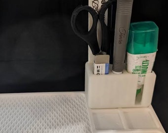 Pode incluir: Um organizador de mesa de plástico branco com uma superfície texturizada e compartimentos. Contém tesoura, um lápis, um estilete, cola e uma borracha. O organizador tem o texto "Da! Da! Moments" num objeto branco.