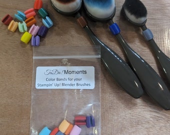 Puede incluir: Una bolsa de plástico transparente con una etiqueta que dice "Ta-Da! Moments Color Bands for your Stampin' Up! Blender Brushes". La bolsa contiene una variedad de gomas elásticas de colores. Tres pinceles mezcladores negros con tapas de diferentes colores son visibles en el fondo.