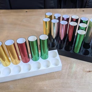 May include: A collection of cylindrical metallic foil rolls in various colors including gold, silver, red, green, and copper. The rolls are organized in two rectangular storage stands, one white and one black, each featuring multiple circular slots.