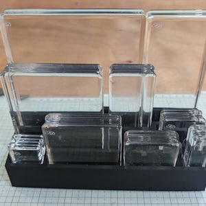May include: A set of clear glass stamp blocks of various sizes, arranged in a black holder. Each block has a rectangular or square shape, with the initials "FD" etched on some. The blocks are designed for crafting and stamping.
