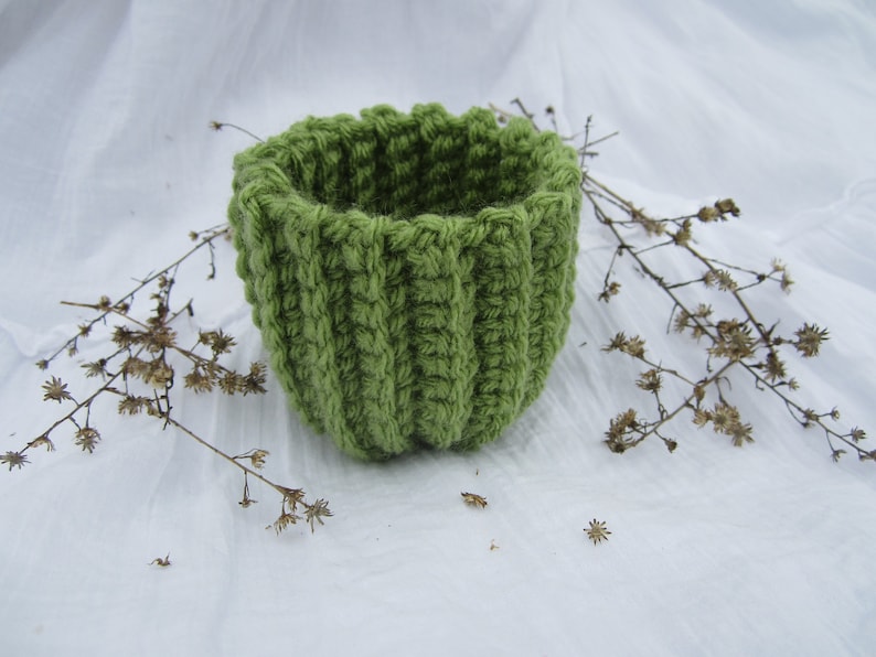May include: A green crocheted basket with a round shape. The basket is made of a soft, textured yarn and has a simple, elegant design.