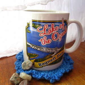 May include: White ceramic mug with a blue and white image of a lake and dam. The mug has the text "Lake of the Ozarks Bagnell Dam" printed on it. The mug is sitting on a blue crocheted coaster.