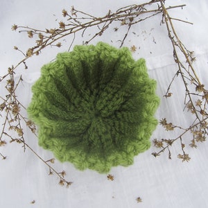 May include: A green crocheted bowl with a textured surface. The bowl is sitting on a white surface.