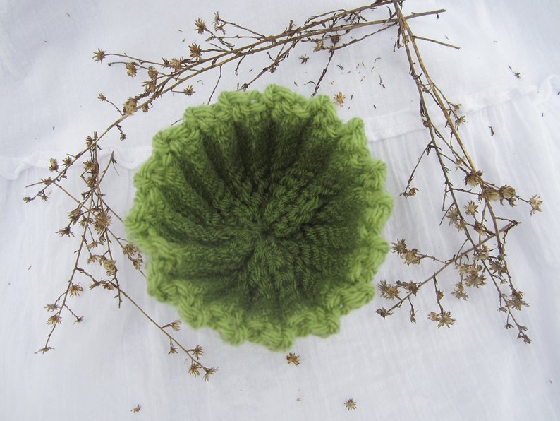 May include: A green crocheted bowl with a textured surface. The bowl is sitting on a white surface.