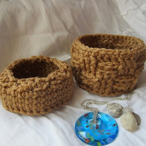 May include: Two brown crocheted baskets of different sizes, one slightly larger than the other, sitting on a white fabric surface. A blue glass pendant with a silver chain is lying on the fabric next to the baskets.