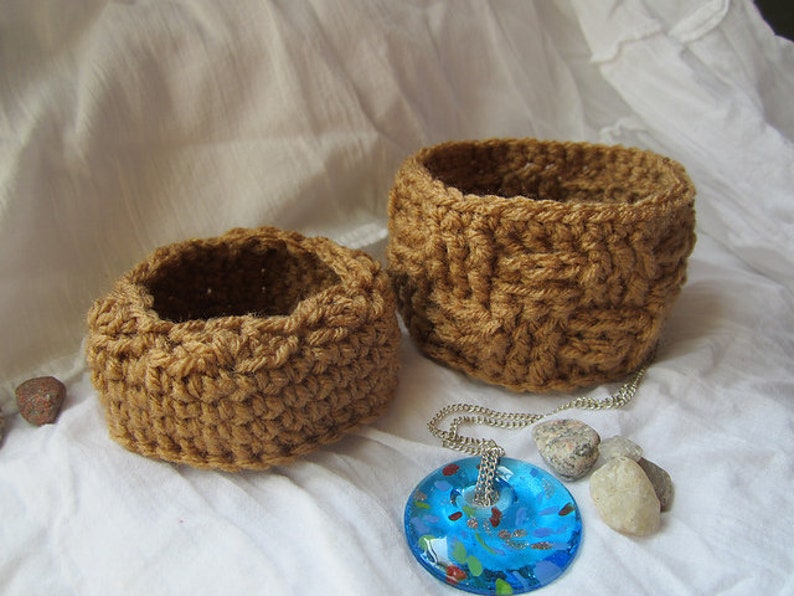 May include: Two brown crocheted baskets of different sizes, one slightly larger than the other, sitting on a white fabric surface. A blue glass pendant with a silver chain is lying on the fabric next to the baskets.
